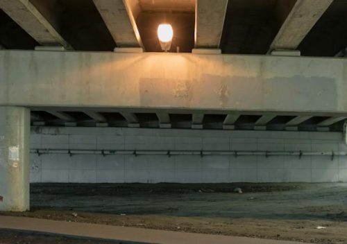 A solitary street lamp glowing under a concrete bridge overpass, casting sharp shadows on the walls and ground.